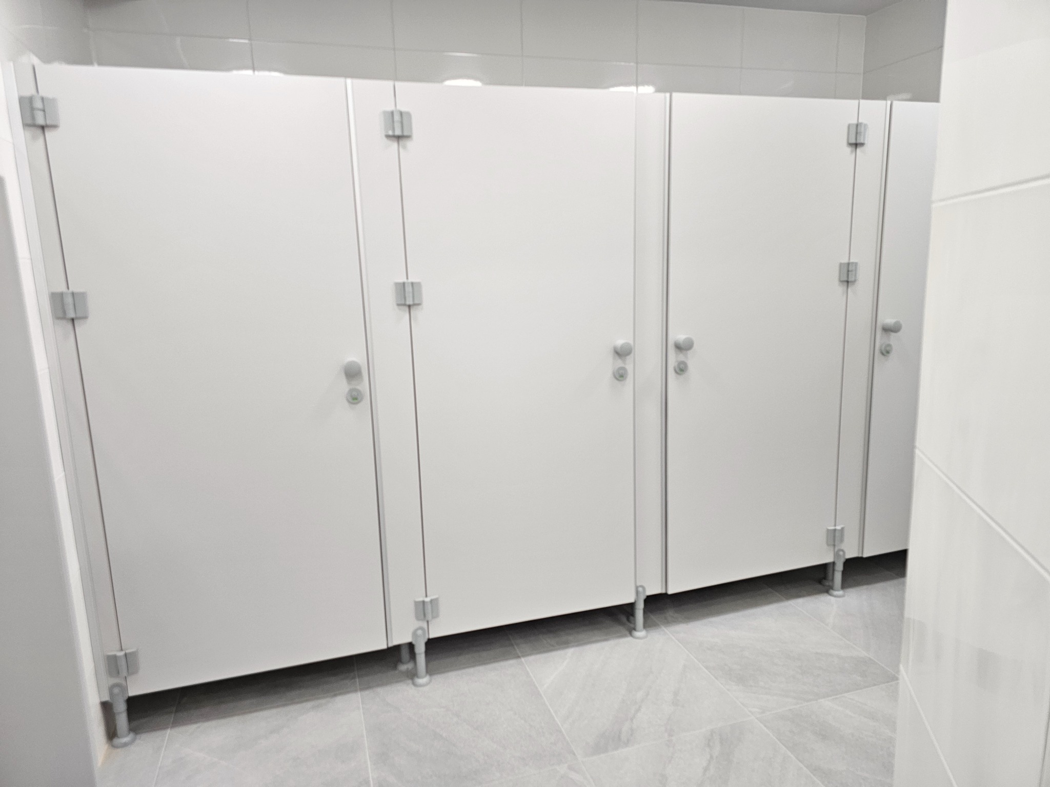 Rząd nowoczesnych, białych kabin WC ze srebrnymi okuciami, klamkami i regulowanymi nóżkami, na tle jasnoszarych płytek podłogowych i białych ściennych, w czystym, minimalistycznym wnętrzu.