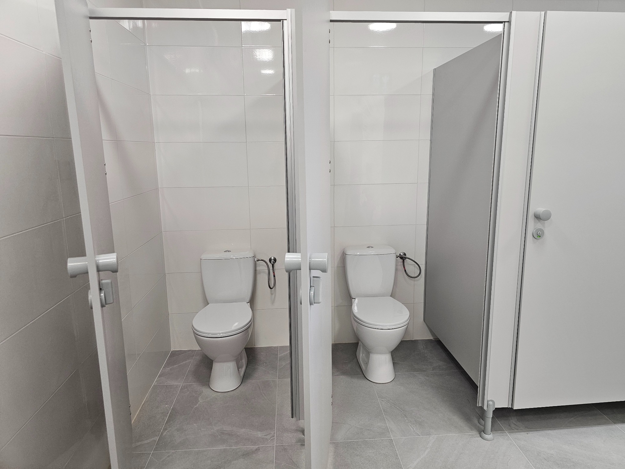 Dwie jasne kabiny toaletowe z białymi kompaktami WC, elastycznymi wężami i szarymi drzwiami, na podłodze szare płytki imitujące marmur. Lewa kabina otwarta.
