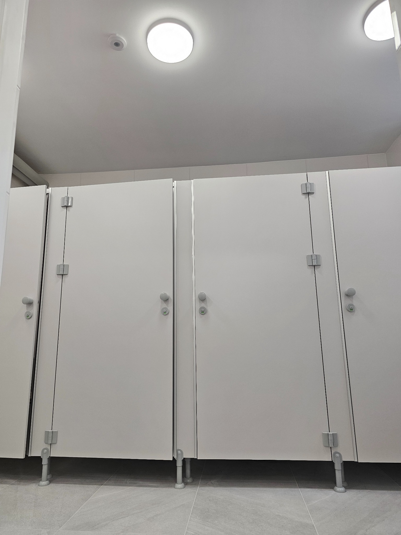 Trzy nowoczesne białe kabiny toaletowe z szarymi uchwytami i zawiasami, na regulowanych nóżkach. Jasne, okrągłe lampy LED na białym suficie, szare płytki podłogowe.