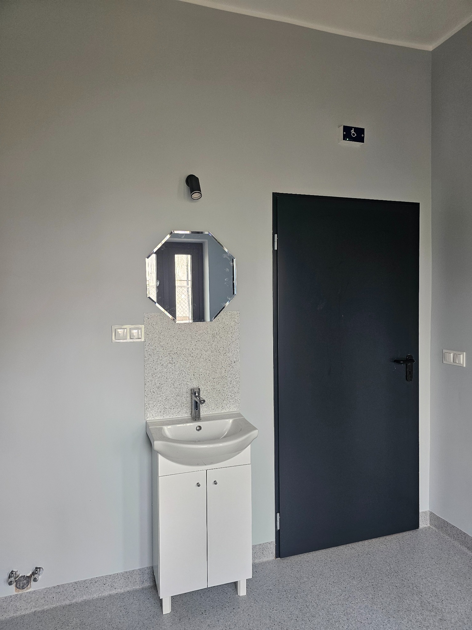 Nowoczesna łazienka z białą umywalką na szafce, ośmiokątnym lustrem i szarymi ścianami. Czarne drzwi i podłoga w kropki, widoczne gniazdka i symbol dostępności.