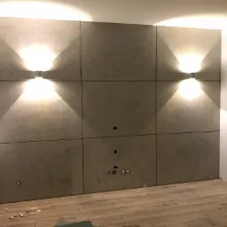 Ściana wykończona panelami z szarego betonu architektonicznego, oświetlona dwoma kinkietami, widoczne gniazdka i otwory na kable, podłoga wyłożona panelami imitującymi drewno.