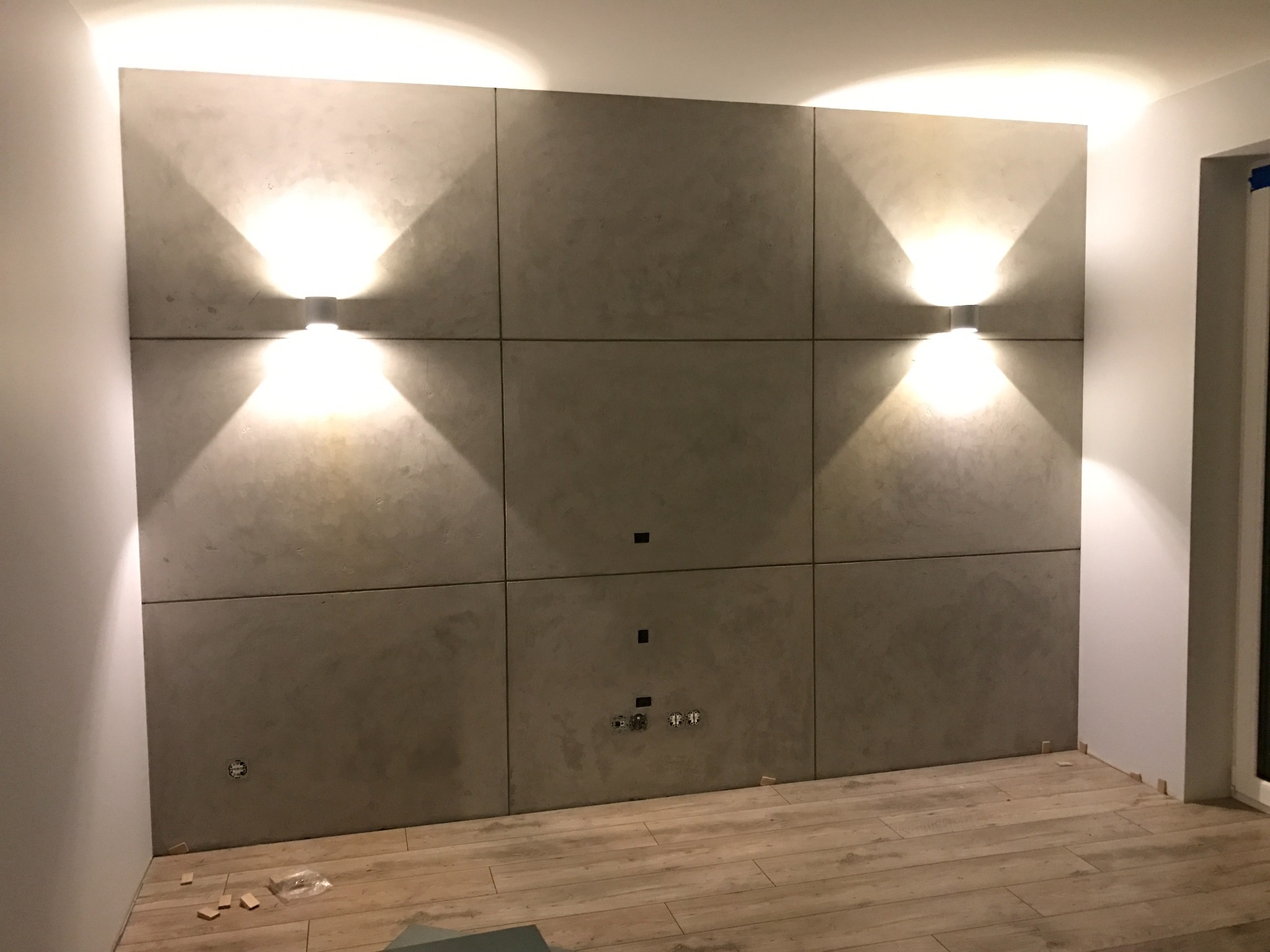 Ściana wykończona panelami z szarego betonu architektonicznego, oświetlona dwoma kinkietami, widoczne gniazdka i otwory na kable, podłoga wyłożona panelami imitującymi drewno.