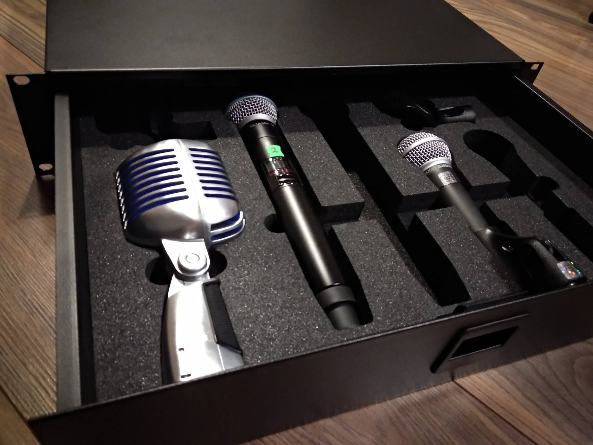 Zestaw trzech mikrofonów w czarnym, wyściełanym pianką pudełku: srebrny mikrofon retro z niebieskimi akcentami, czarny mikrofon bezprzewodowy z zieloną naklejką i srebrny mikrofon dynamiczny Shure...