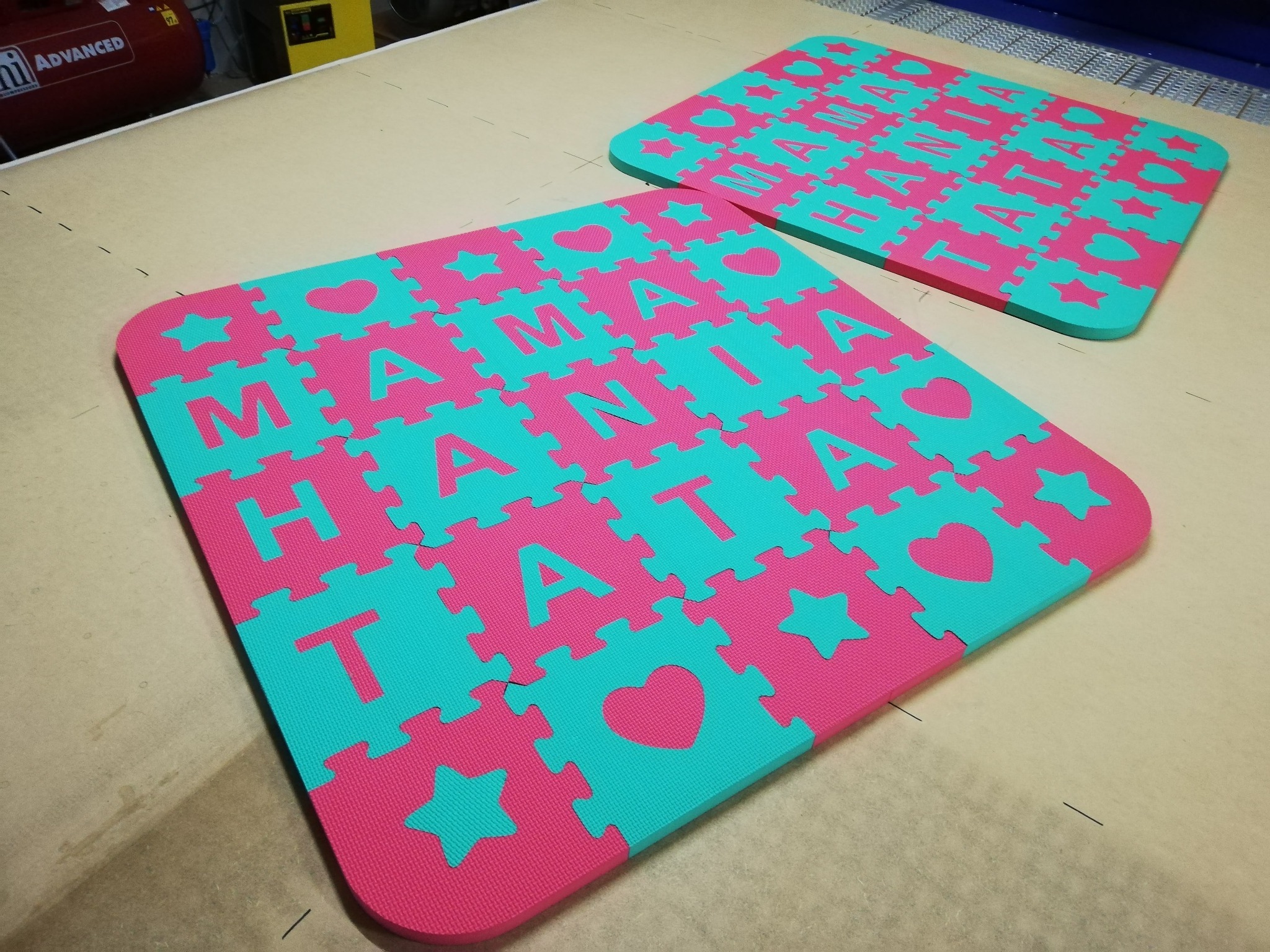 Dwie kwadratowe maty piankowe puzzle w kolorach różowym i turkusowym, z wyciętymi kształtami liter, gwiazd i serc, ułożone na beżowej powierzchni.