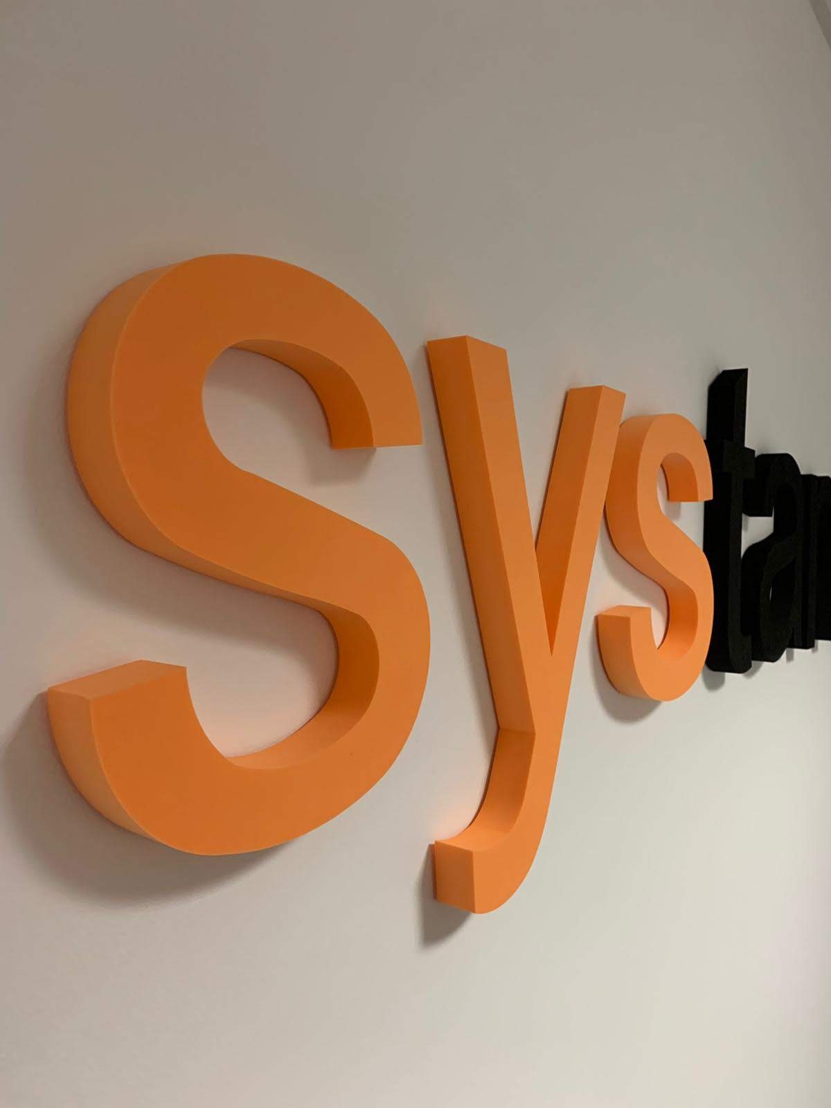 Pomarańczowe i czarne trójwymiarowe litery tworzące napis na białej ścianie, widoczne pod kątem.