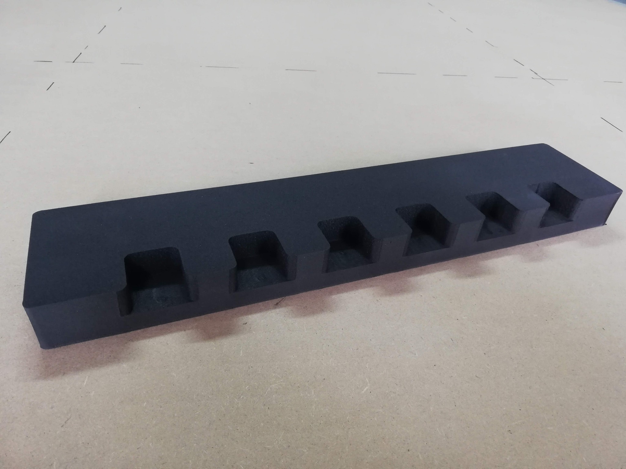 Czarny, prostokątny wkład z pianki z wyciętymi wgłębieniami, leżący na beżowym tle, prawdopodobnie element opakowania ochronnego.