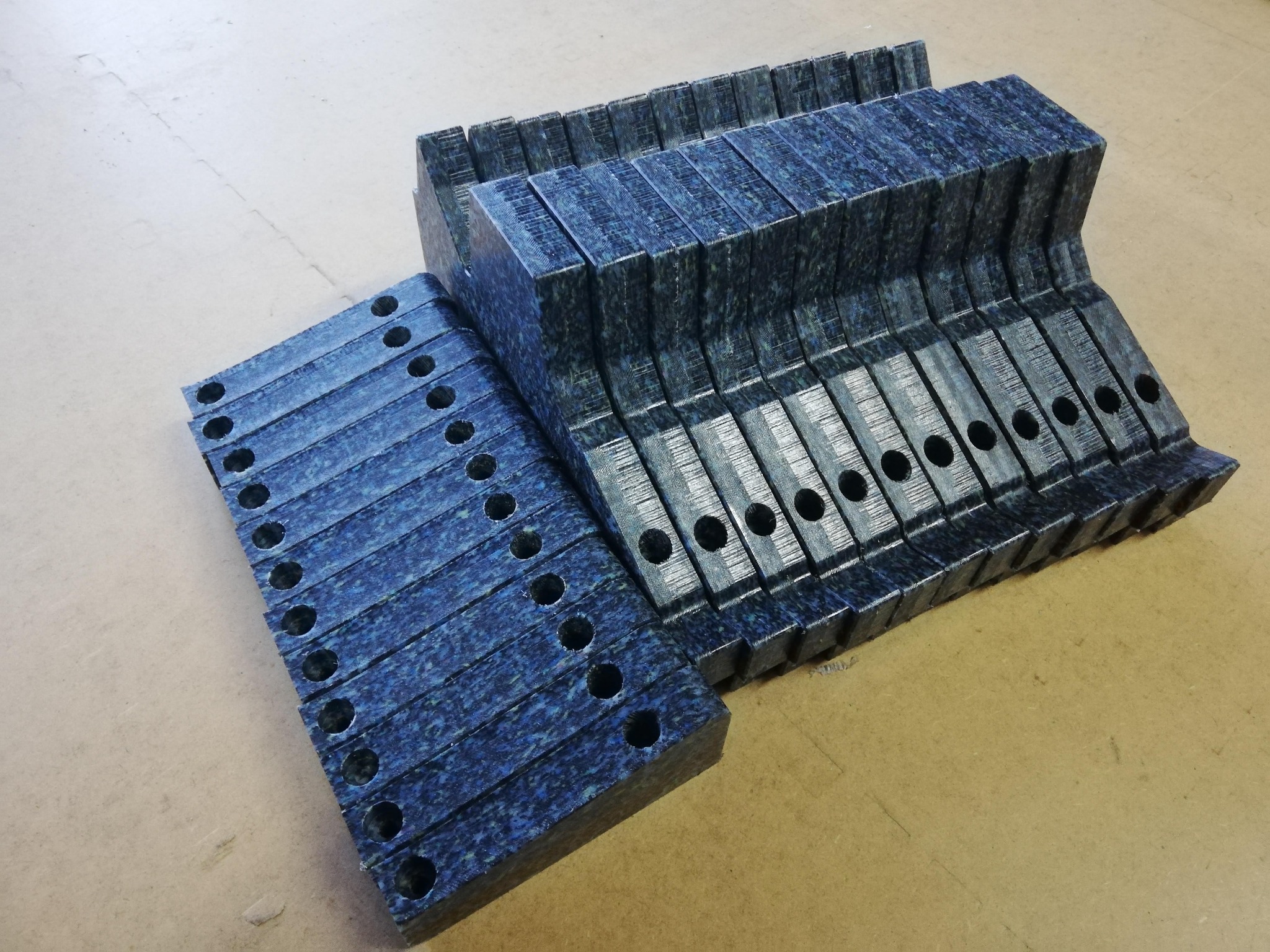 Stos ciemnych, niebiesko-czarnych elementów z tworzywa sztucznego z otworami, ułożonych w dwóch grupach na jasnym tle, prawdopodobnie przygotowanych do pakowania lub transportu.