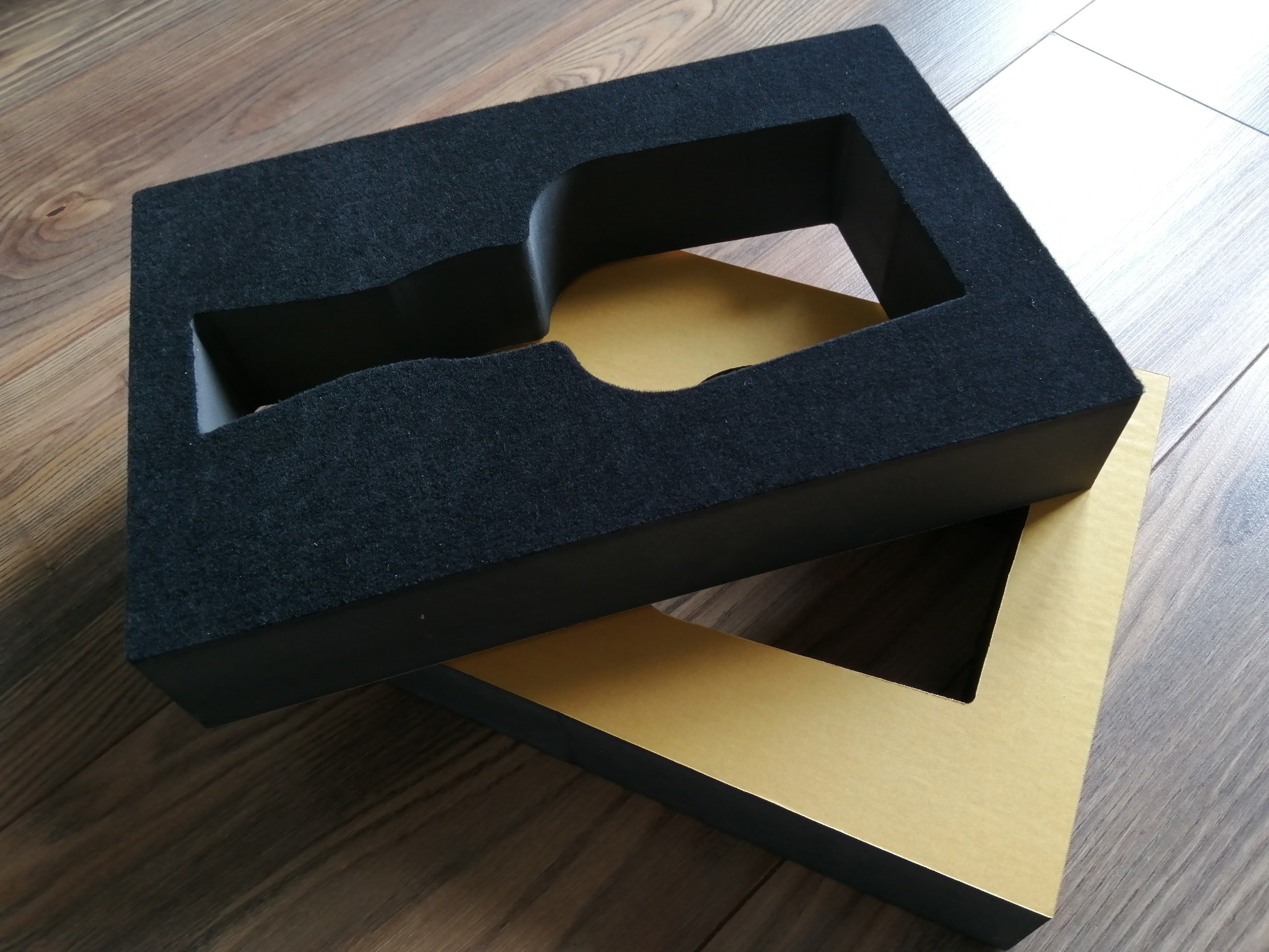 Dwa pudełka: czarne z gąbką z wyciętym kształtem i złote, leżące jedno na drugim na drewnianej podłodze.