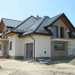 Nowoczesny dom jednorodzinny z ciemnoszarą dachówką, jasną elewacją z kamienną podmurówką i drewnianymi elementami wykończeniowymi, widoczny w słoneczny dzień z placem budowy w tle.