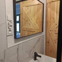Nowoczesna łazienka z umywalką, czarną baterią i lustrem z drewnianą ramą, odbijającym drewniane drzwi przesuwne typu stodoła.