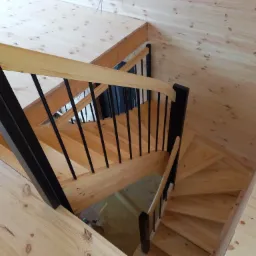 Wewnętrzne spiralne schody z jasnego drewna z czarnymi metalowymi prętami balustrady, widok z góry na drewniane wykończenie ścian i podłogi.