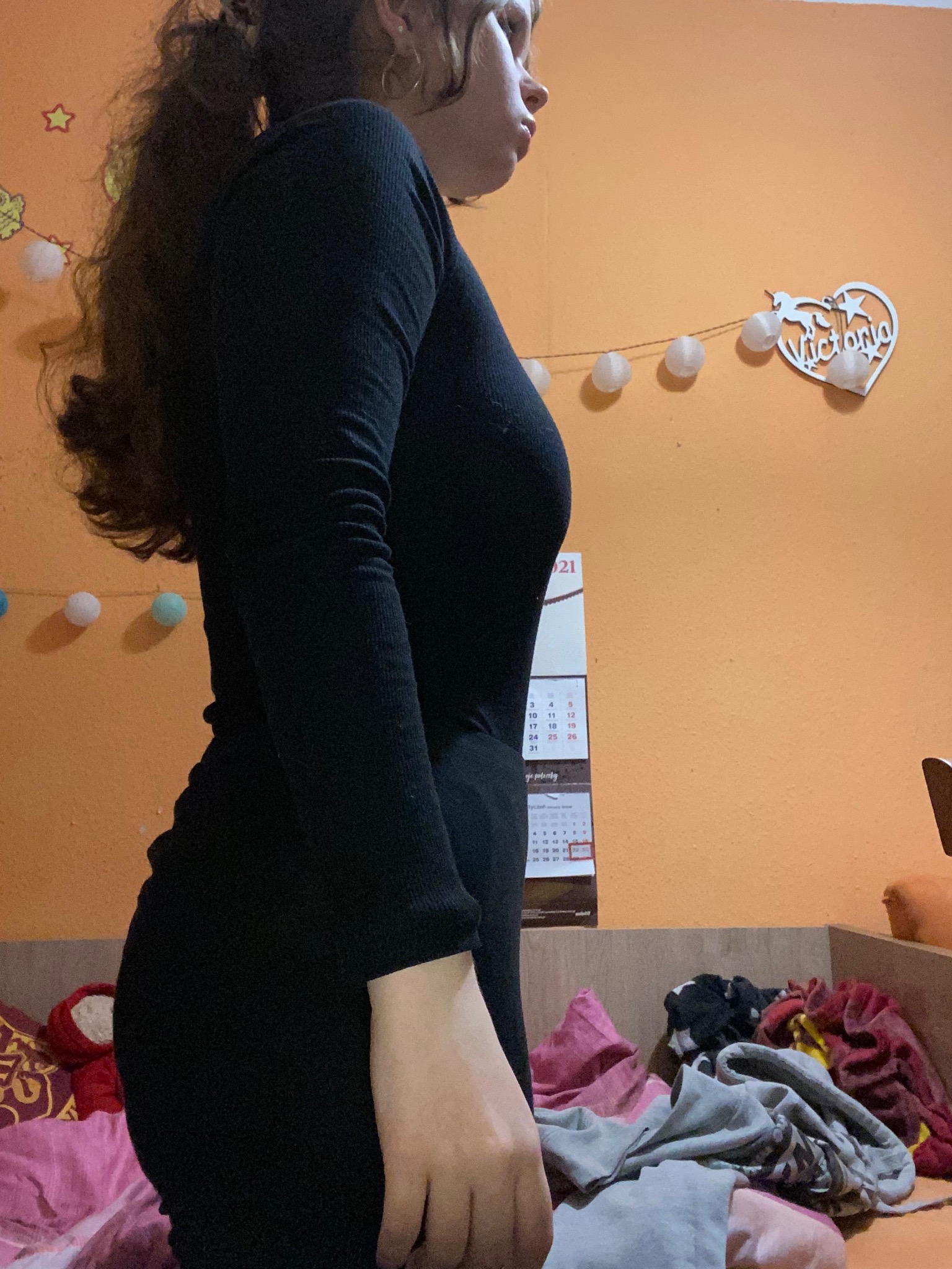 Kobieta w czarnym obcisłym stroju, stojąca bokiem na tle pomarańczowej ściany z kalendarzem i dekoracją w kształcie serca z napisem Victoria.