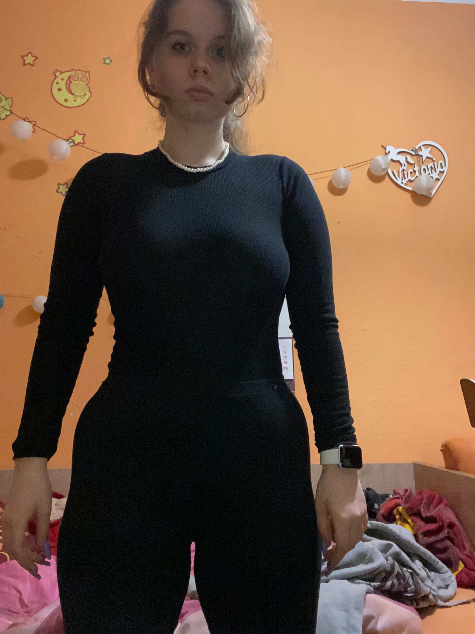 Portret młodej kobiety w czarnym kombinezonie na tle pomarańczowej ściany z dekoracjami, widoczny zegarek na ręce i perłowy naszyjnik.