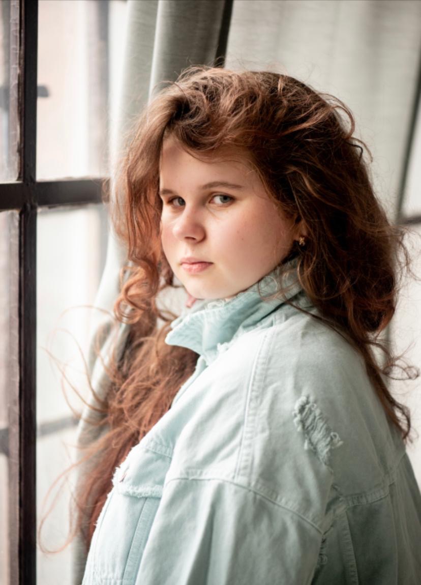 Portret młodej kobiety z długimi, rudymi włosami, ubrana w jasną kurtkę dżinsową, spogląda przez ramię w stronę obiektywu, stojąc przy oknie z ciemną ramą.