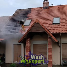 Wash Expert - Częściowo wyczyszczony dach z czerwonej dachówki. Pracownik w kasku czyści ciemny nalot. Widoczny budynek z ceglanymi kolumnami i logo firmy.