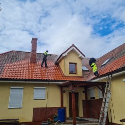 Wash Expert - Dwóch pracowników w jaskrawych strojach czyści dach z czerwonej dachówki za pomocą myjki ciśnieniowej. Widoczna drabina, żółta elewacja domu i błękitne niebo.