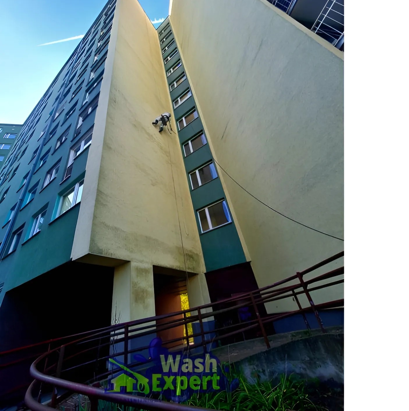 Pracownik na linie czyści elewację wysokiego budynku. Widok z dołu, z rampą i zielenią w kadrze. Częściowo zabrudzona ściana przed czyszczeniem.