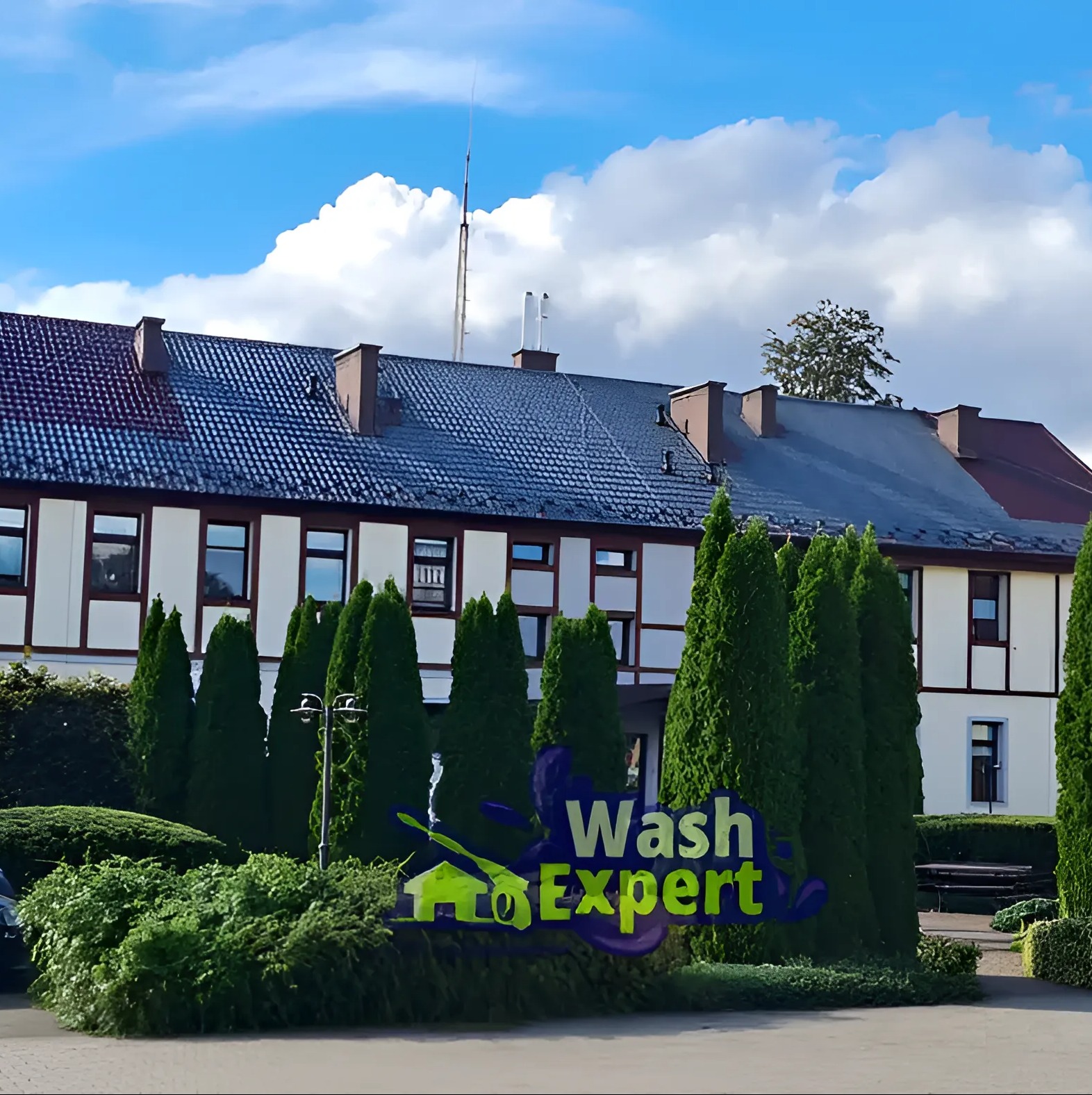 Budynek z białymi ścianami i ciemnym dachem, otoczony zielenią. Widoczne logo firmy 'Wash Expert'.