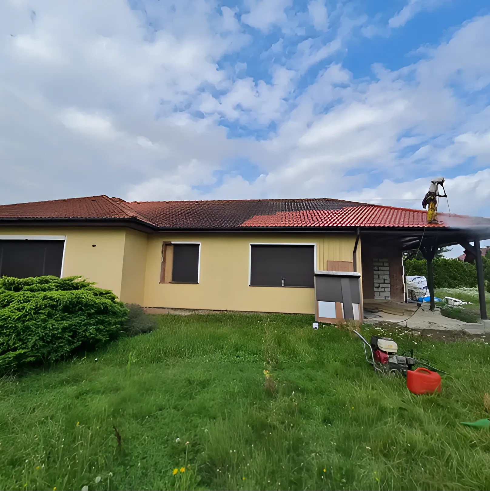 Pracownik w trakcie mycia dachu z czerwonej dachówki. Część dachu jest już umyta, ukazując różnicę w kolorze. W tle żółta elewacja domu i błękitne niebo.