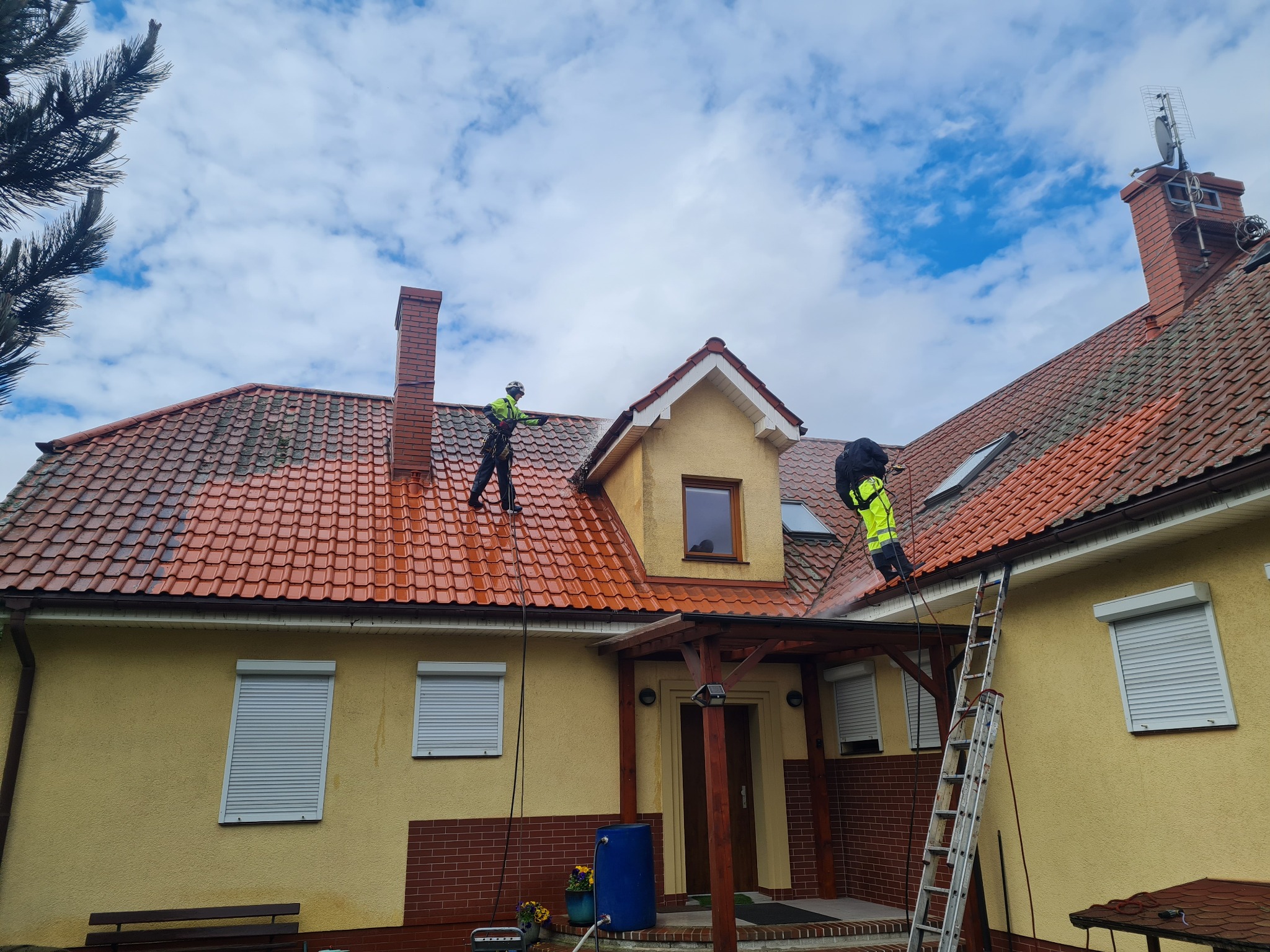 Dwóch pracowników w jaskrawych strojach czyści dach z czerwonej dachówki za pomocą myjki ciśnieniowej. Widoczna drabina, żółta elewacja domu i błękitne niebo.