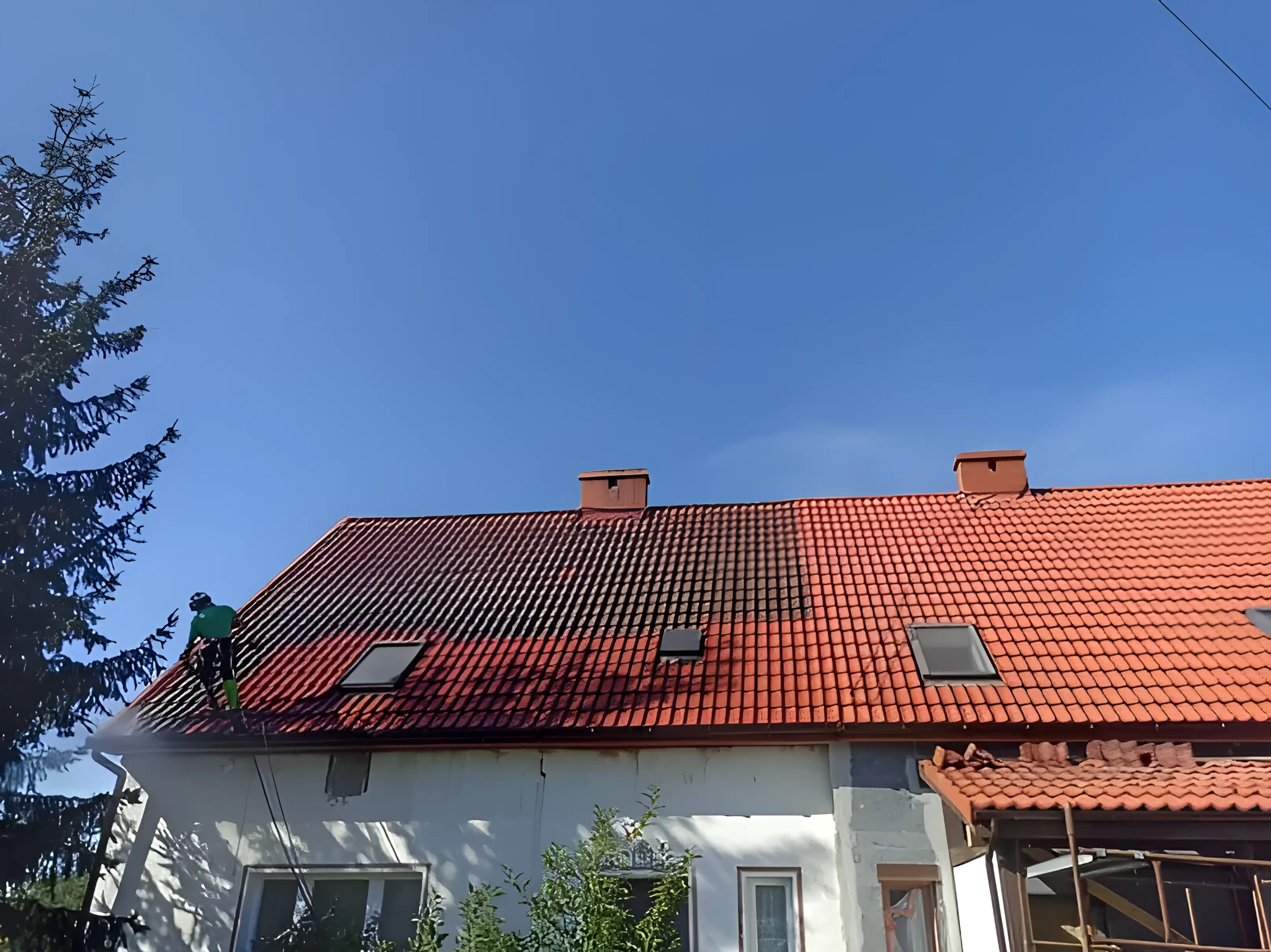 Częściowo wyczyszczony dach z czerwonej dachówki podczas pracy. Widoczna różnica w kolorze między czystą a brudną częścią dachu. Pracownik w zielonym kombinezonie.