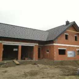 Dom w trakcie budowy z ciemną dachówką i widocznymi pustakami. Na ścianie baner reklamowy. Widoczne rynny. Surowy stan budynku.