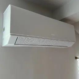 Biała klimatyzacja AUX, zamontowana na szarej ścianie w pomieszczeniu. Widoczna kratka wylotowa i logo producenta. Ujęcie z dołu, lekko z prawej strony.