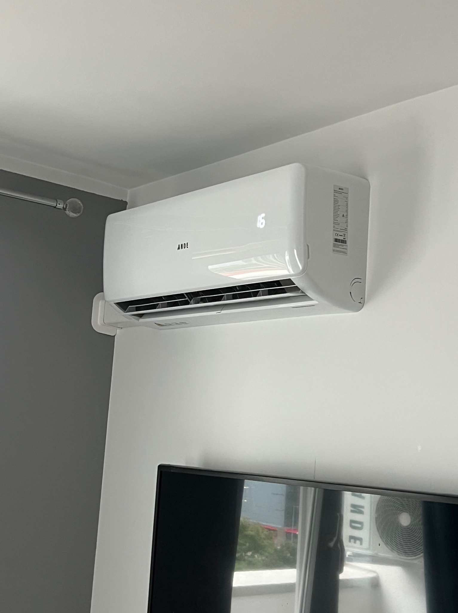 Biały klimatyzator marki Ande zamontowany na ścianie, z wyświetlaczem pokazującym temperaturę 16 stopni, widoczny fragment telewizora w dolnej części kadru.