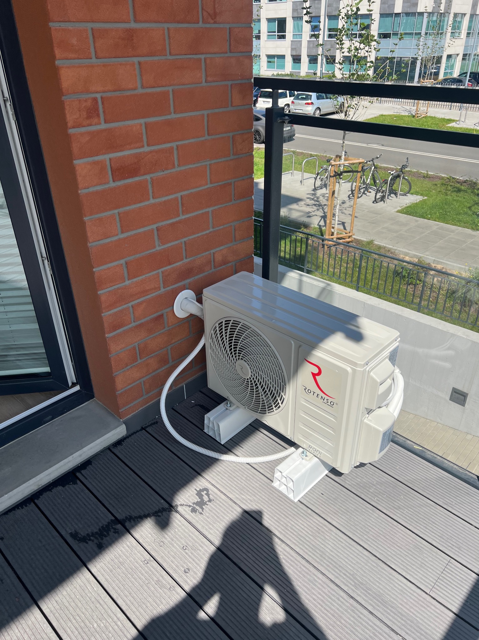 Klimatyzator Rotenso zamontowany na balkonie z deską kompozytową, widoczny fragment ceglanej ściany i budynki w tle. Czysty montaż.