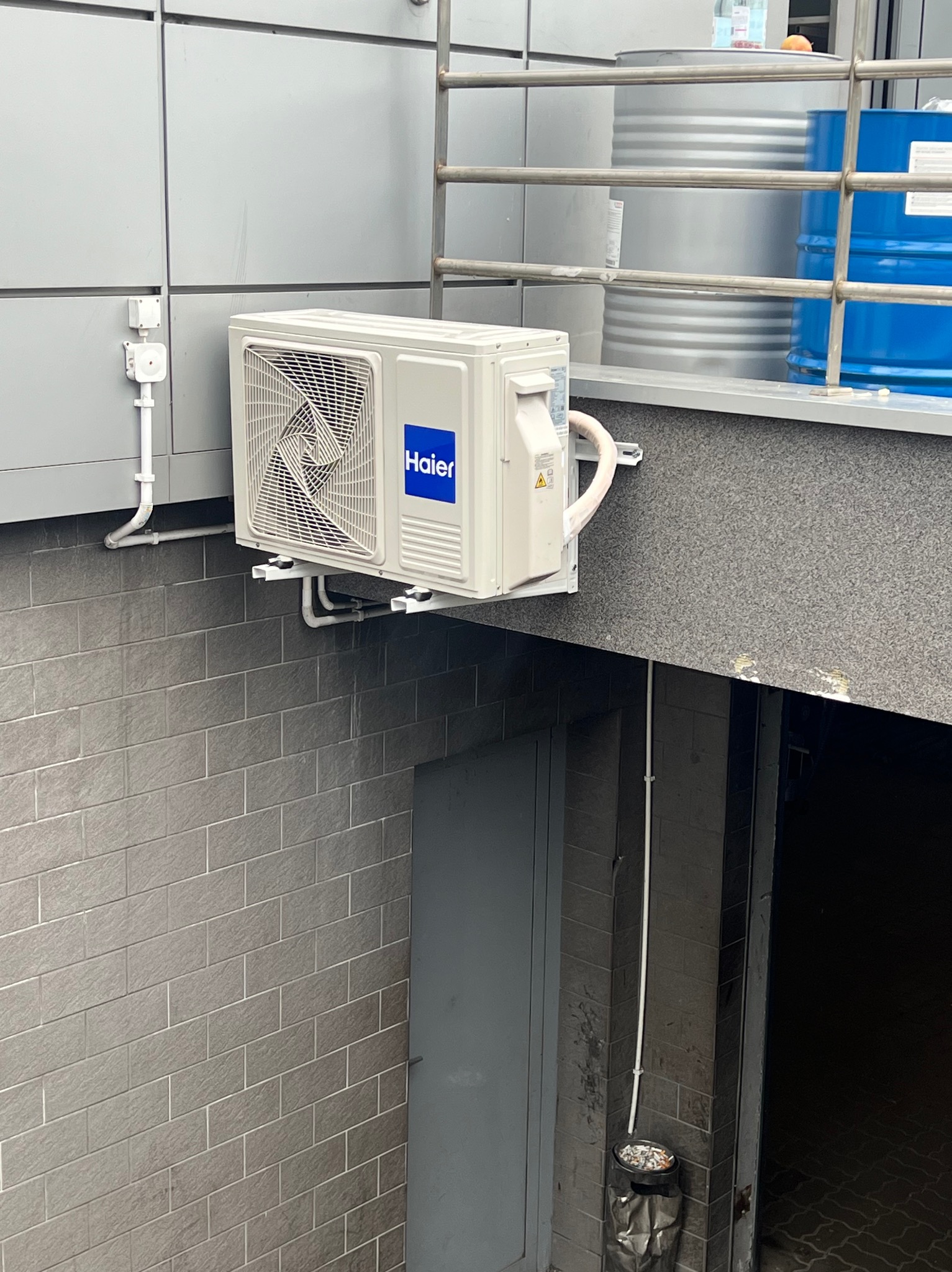 Klimatyzator Haier zamontowany na szarej ścianie budynku z widocznymi elementami instalacji. W tle metalowe beczki i fragment konstrukcji. Widoczny kosz na śmieci.