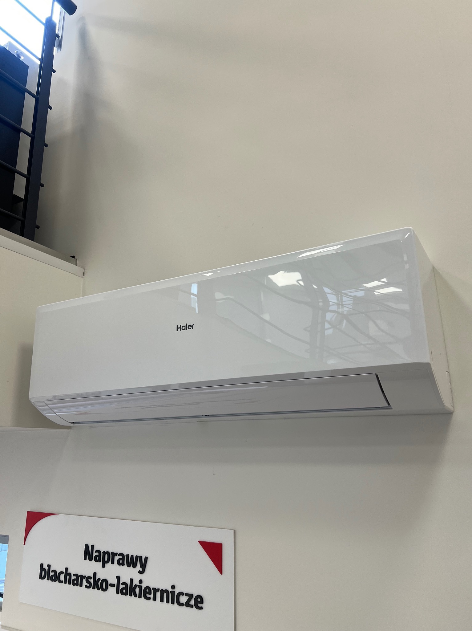 Biały klimatyzator Haier na ścianie, widoczny fragment schodów i szyld warsztatu blacharsko-lakierniczego. Minimalistyczny design, jasne wnętrze.