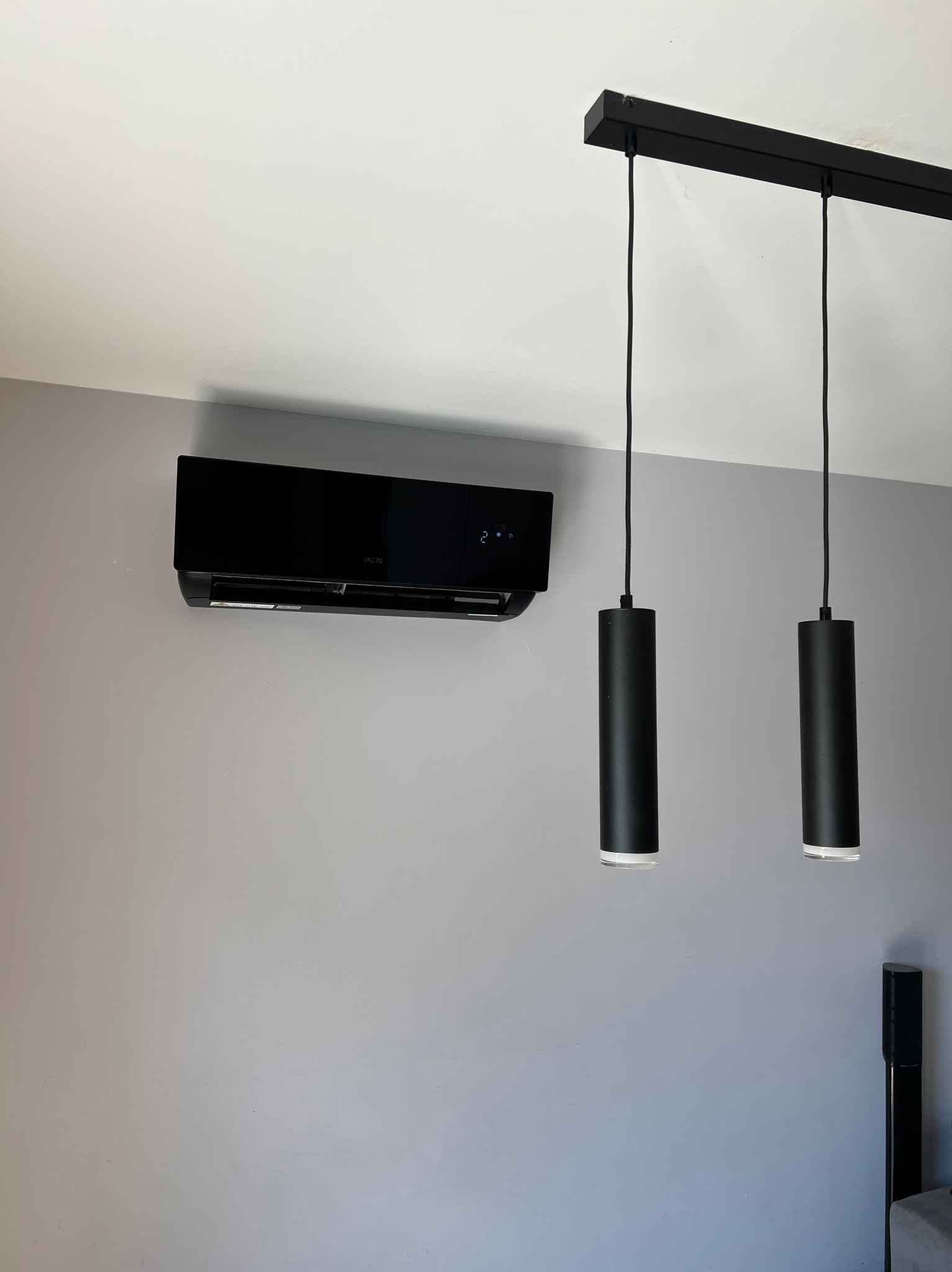 Elegancki, czarny klimatyzator ścienny marki AUX, zamontowany w nowoczesnym wnętrzu z szarą ścianą i czarnymi lampami wiszącymi. Widoczne cyfry na wyświetlaczu.