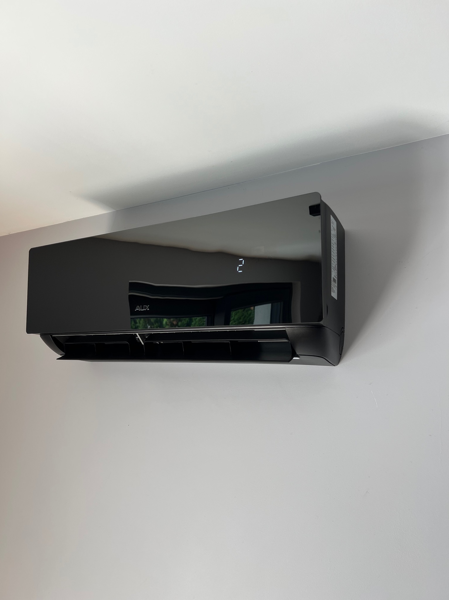 Czarna, błyszcząca klimatyzacja AUX na ścianie, z widocznym wyświetlaczem temperatury i odbiciem okna w obudowie, zamontowana w jasnym pomieszczeniu.