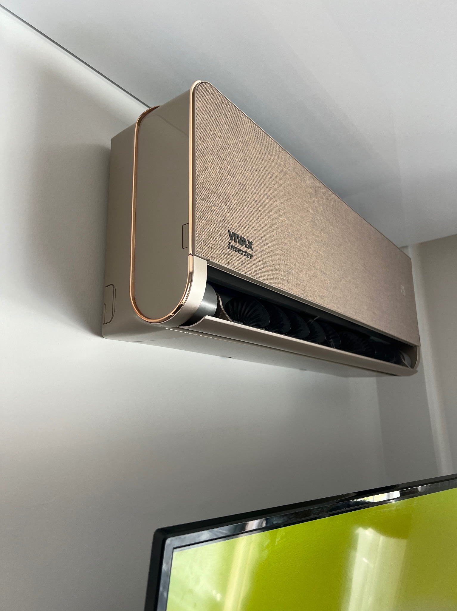 Beżowy klimatyzator VIVAX Inverter z miedzianymi akcentami, zamontowany na białej ścianie w pomieszczeniu. Widoczny fragment telewizora w dolnej części kadru.