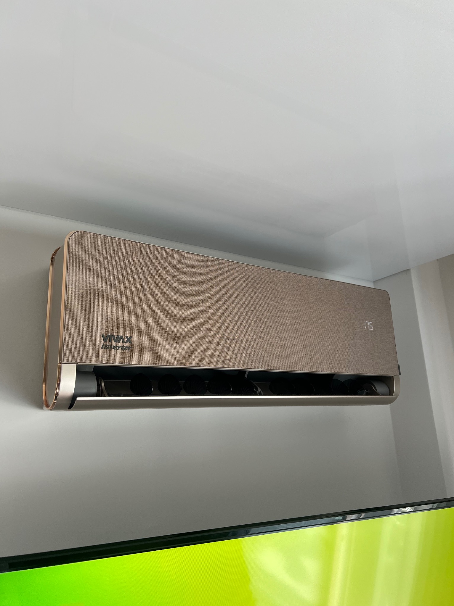 Klimatyzator Vivax Inverter o teksturowanej, złotej obudowie, zamontowany na ścianie w pomieszczeniu. Widoczny fragment telewizora w dolnej części kadru.