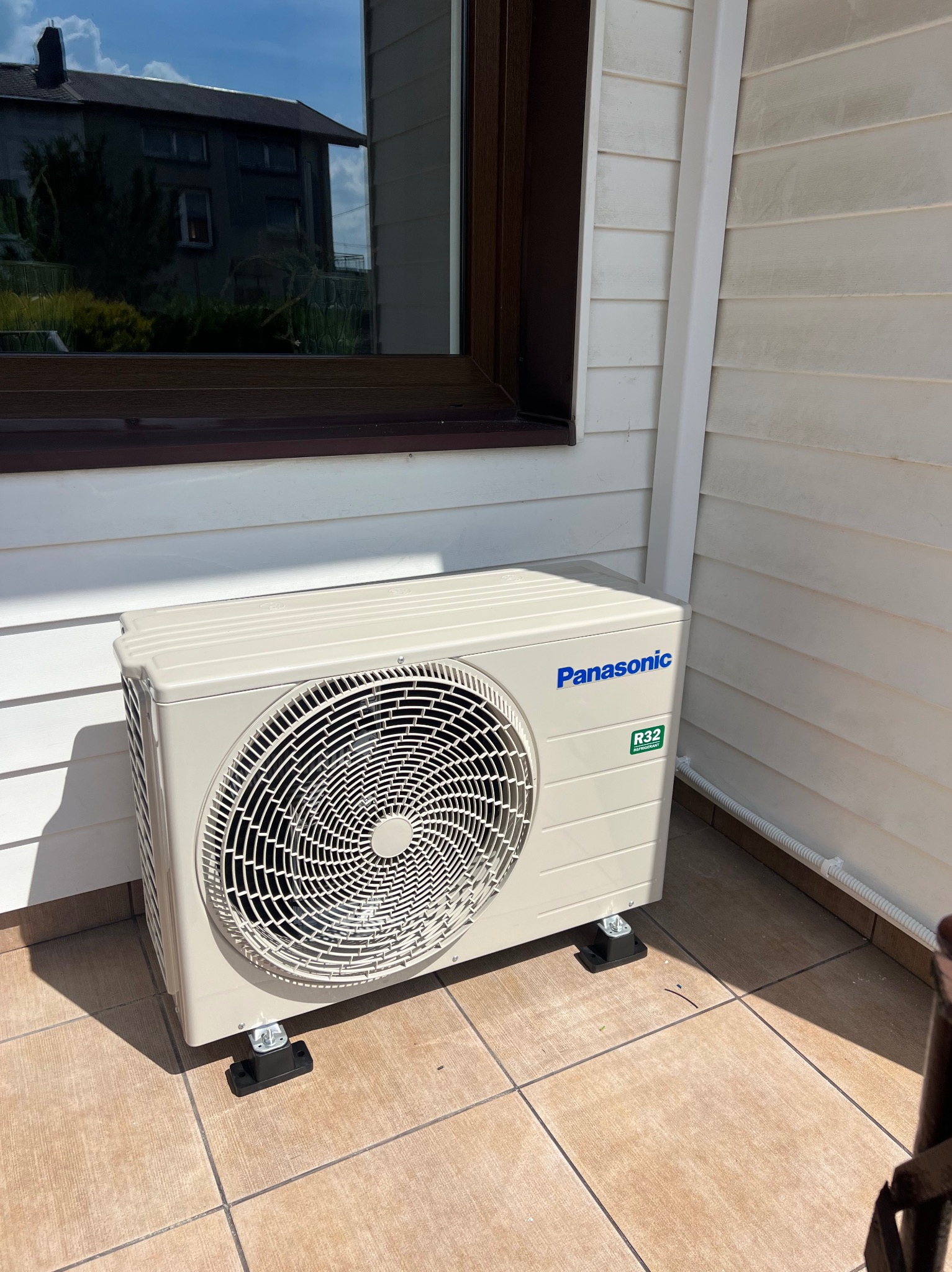 Zewnętrzna jednostka klimatyzacji Panasonic zamontowana na wspornikach przy ścianie budynku z oknem. Widoczne płytki na podłożu i biała elewacja.