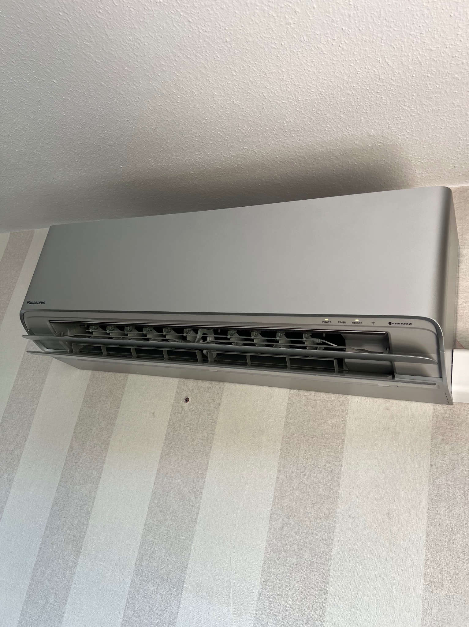 Zamontowana klimatyzacja Panasonic na ścianie w paski. Widoczny otwarty panel przedni i wlot powietrza. Oświetlenie naturalne, wnętrze mieszkalne.