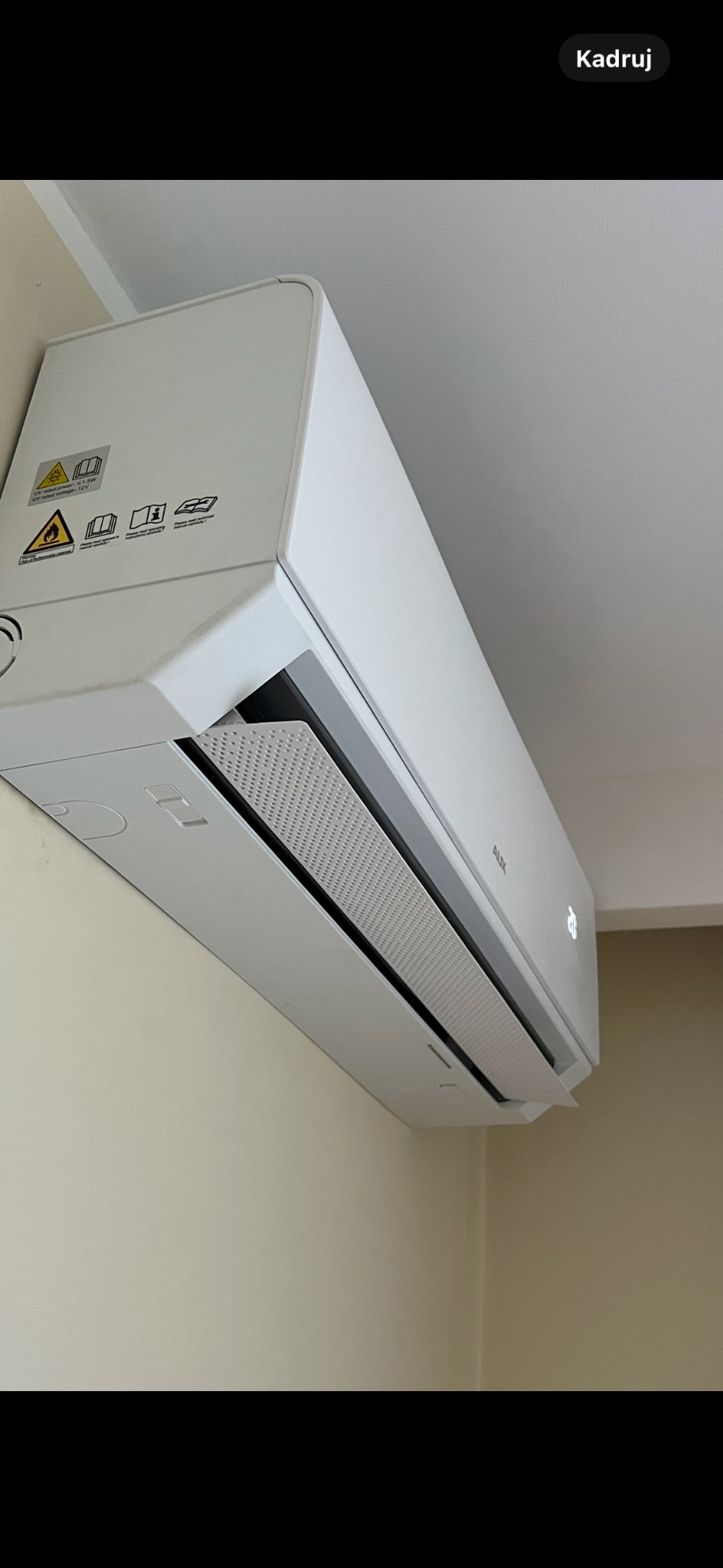 Biały klimatyzator marki AUX zamontowany na ścianie w jasnym pomieszczeniu. Widoczny panel z kratką wentylacyjną i wyświetlacz temperatury.