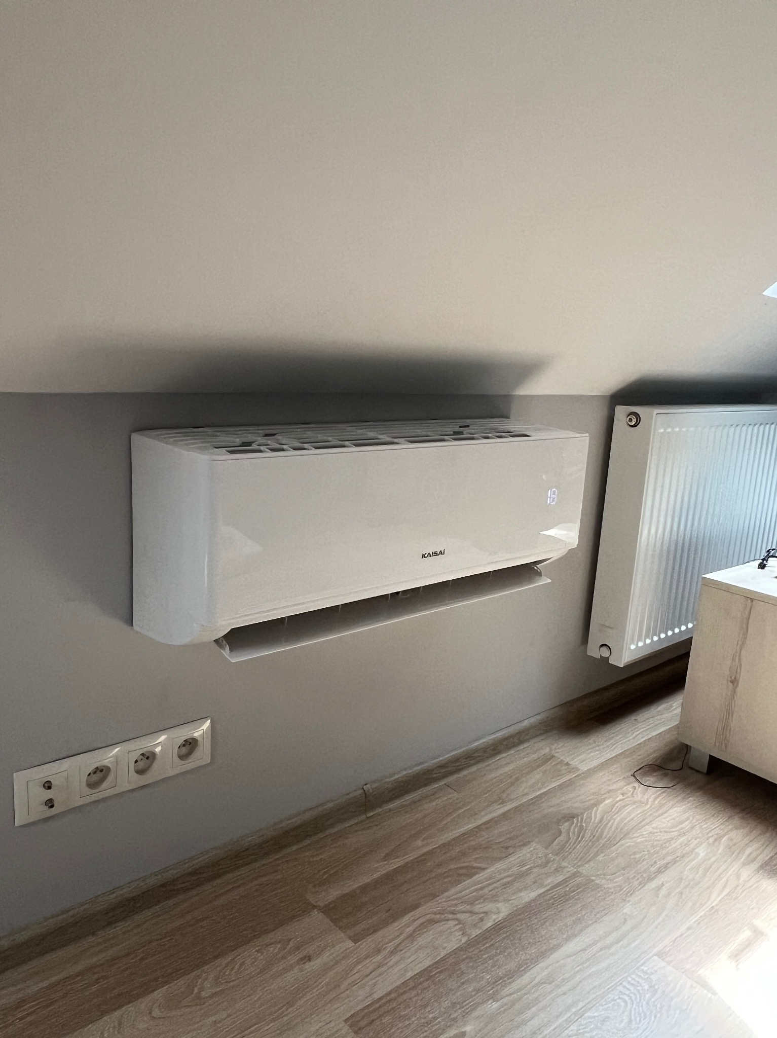 Biały klimatyzator KaisaI na szarej ścianie pod skosem, obok grzejnika i gniazdek. Wyświetla temperaturę 18 stopni. Podłoga panelowa.