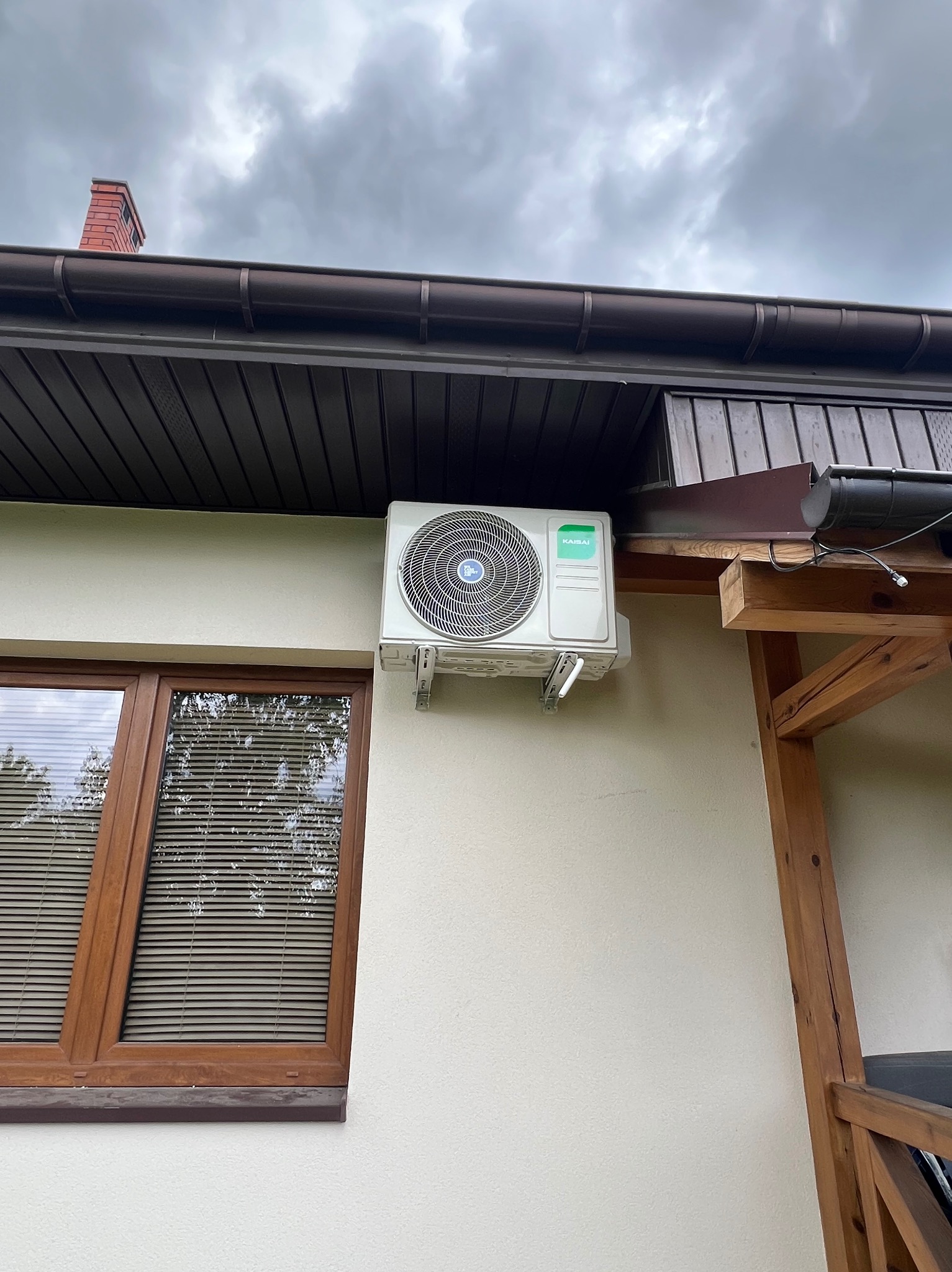 Biała klimatyzacja marki KAISAI zamontowana na elewacji domu, widoczna obok okna z brązową ramą i drewnianej konstrukcji. W tle pochmurne niebo.