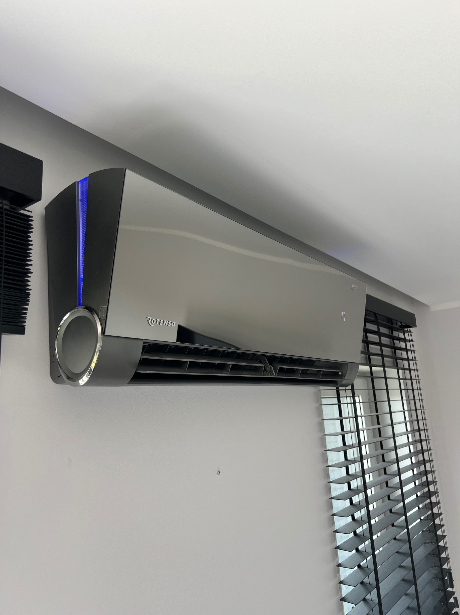 Nowoczesny klimatyzator Rotenso w kolorze srebrnym, zamontowany na białej ścianie, z widocznymi czarnymi żaluzjami w tle. Urządzenie z podświetleniem LED.