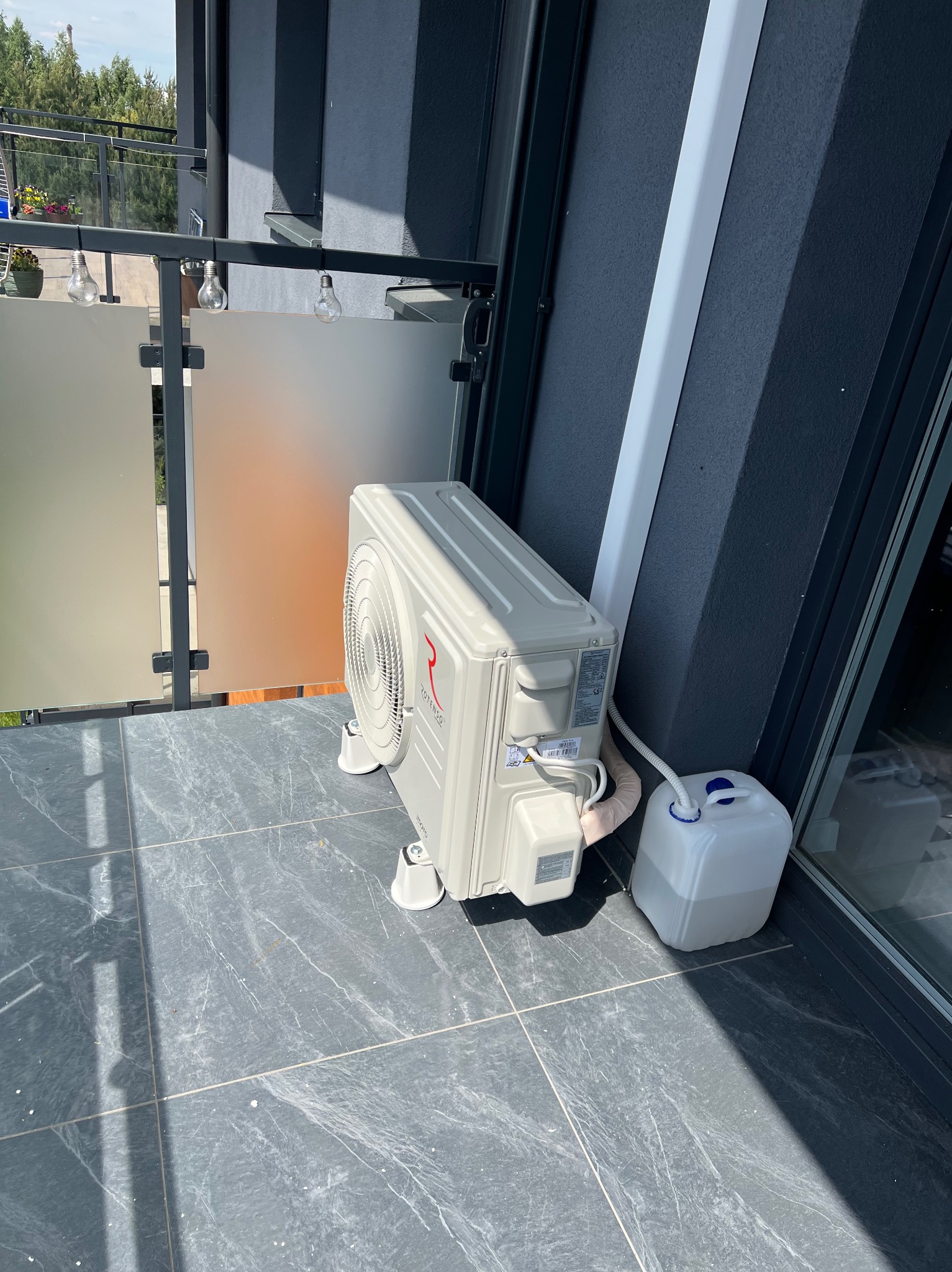 Biały klimatyzator Rotenso na balkonie z szarymi płytkami, obok biały pojemnik na wodę. Widoczne elementy instalacji i nowoczesna balustrada.