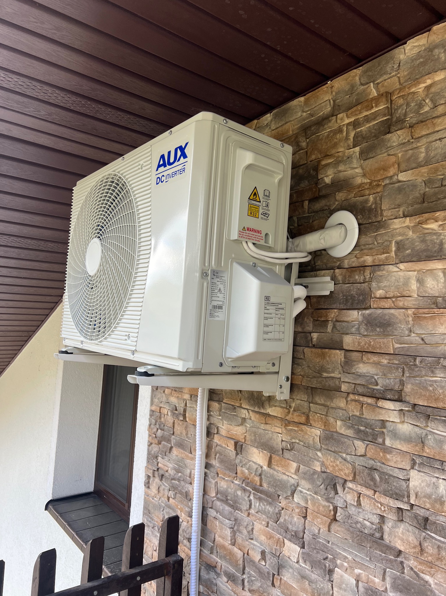 Klimatyzator AUX DC Inverter zamontowany na ścianie z kamienia dekoracyjnego, widoczne brązowe elementy wykończenia dachu i okna. Instalacja w domu.