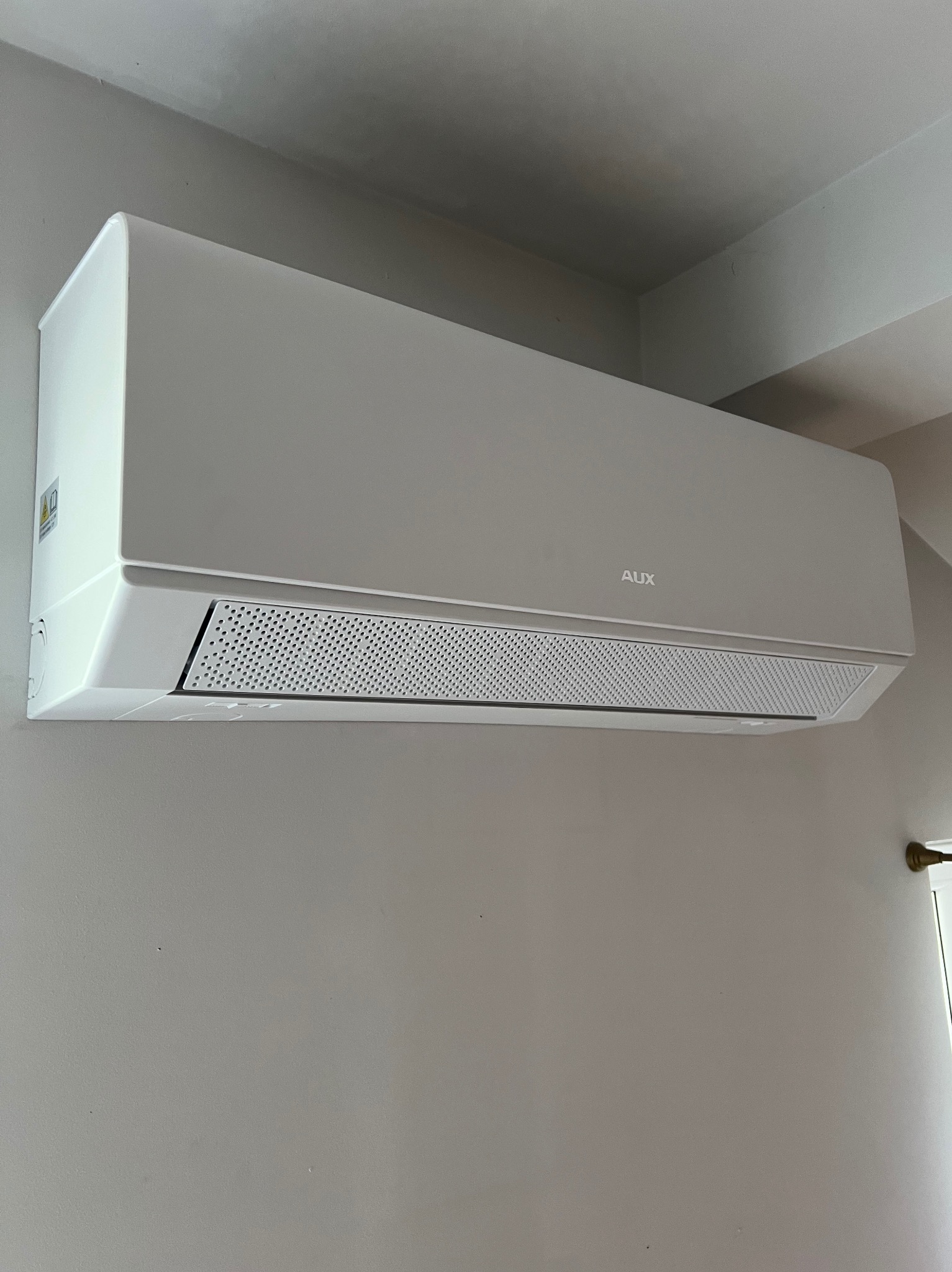 Biała klimatyzacja AUX, zamontowana na szarej ścianie w pomieszczeniu. Widoczna kratka wylotowa i logo producenta. Ujęcie z dołu, lekko z prawej strony.