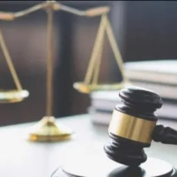 Złota waga sprawiedliwości i drewniany młotek sędziowski na tle stosu książek prawniczych na białym stole.