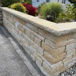 Narożnik ogrodzenia z piaskowca, widok z poziomu chodnika, w tle roślinność ogrodowa i dom.