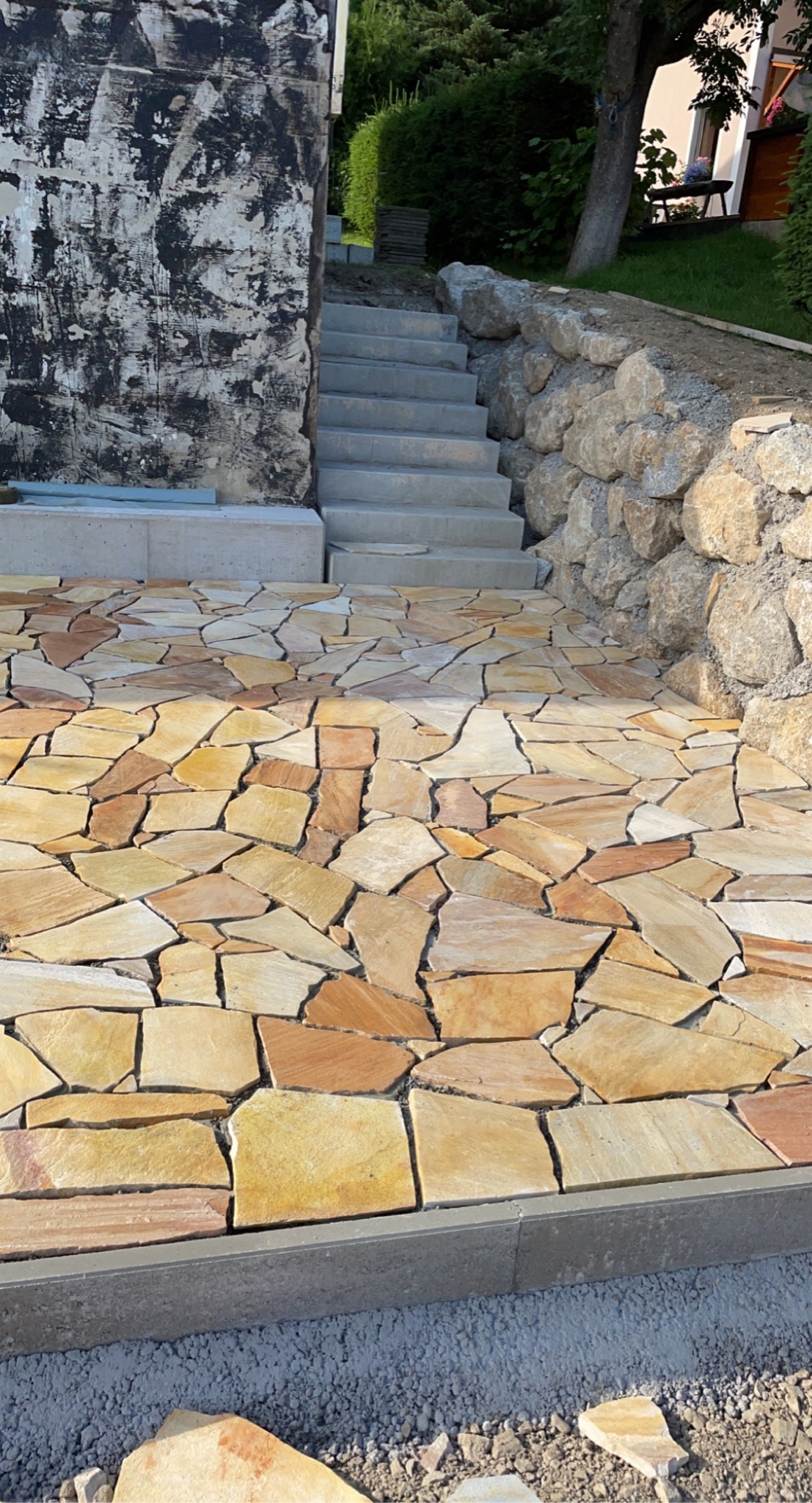 Ułożony z nieregularnych, żółto-brązowych płyt kamiennych taras przechodzi w betonowe schody prowadzące w górę, obok kamienny murek oporowy.