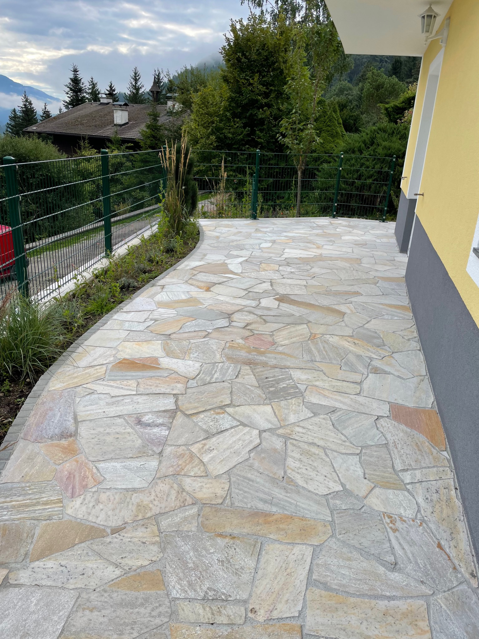 Wykończony taras z nieregularnych płyt kamiennych w odcieniach szarości i beżu, otoczony zielonym metalowym ogrodzeniem i rabatami z trawami ozdobnymi, z widokiem na zalesione wzgórza w tle.