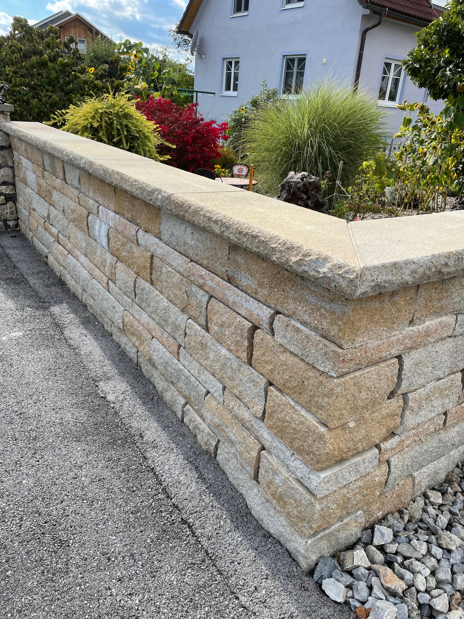 Narożnik ogrodzenia z piaskowca, widok z poziomu chodnika, w tle roślinność ogrodowa i dom.