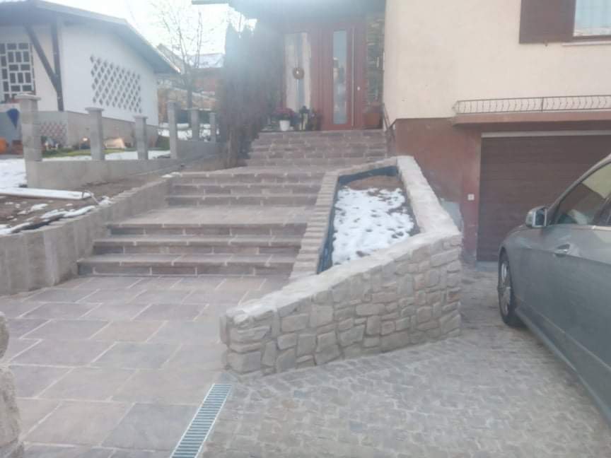 Kamienne schody prowadzące do domu z donicą obok, wyłożony kamienną kostką podjazd i częściowo ośnieżony teren.
